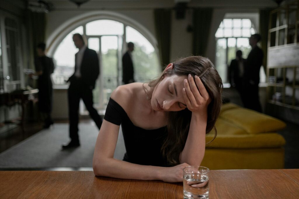 A stressed woman in a formal indoor setting, with a glass of water and blurred figures in the background.