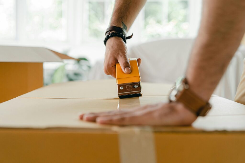 Close-up of a man sealing a cardboard box with a tape dispenser during a move indoors.